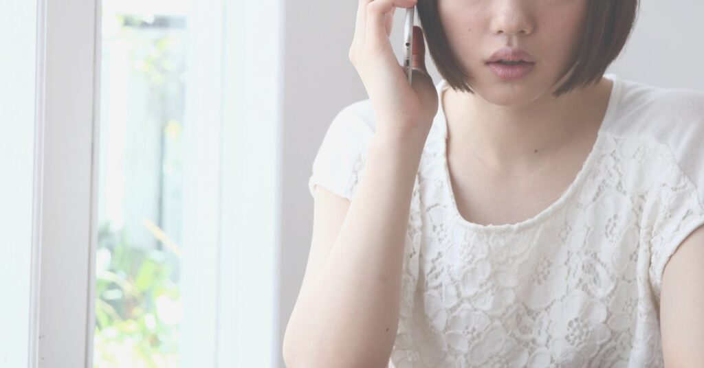 窓辺で支援先に電話する女性の横顔のシルエットとメモを取る手元。